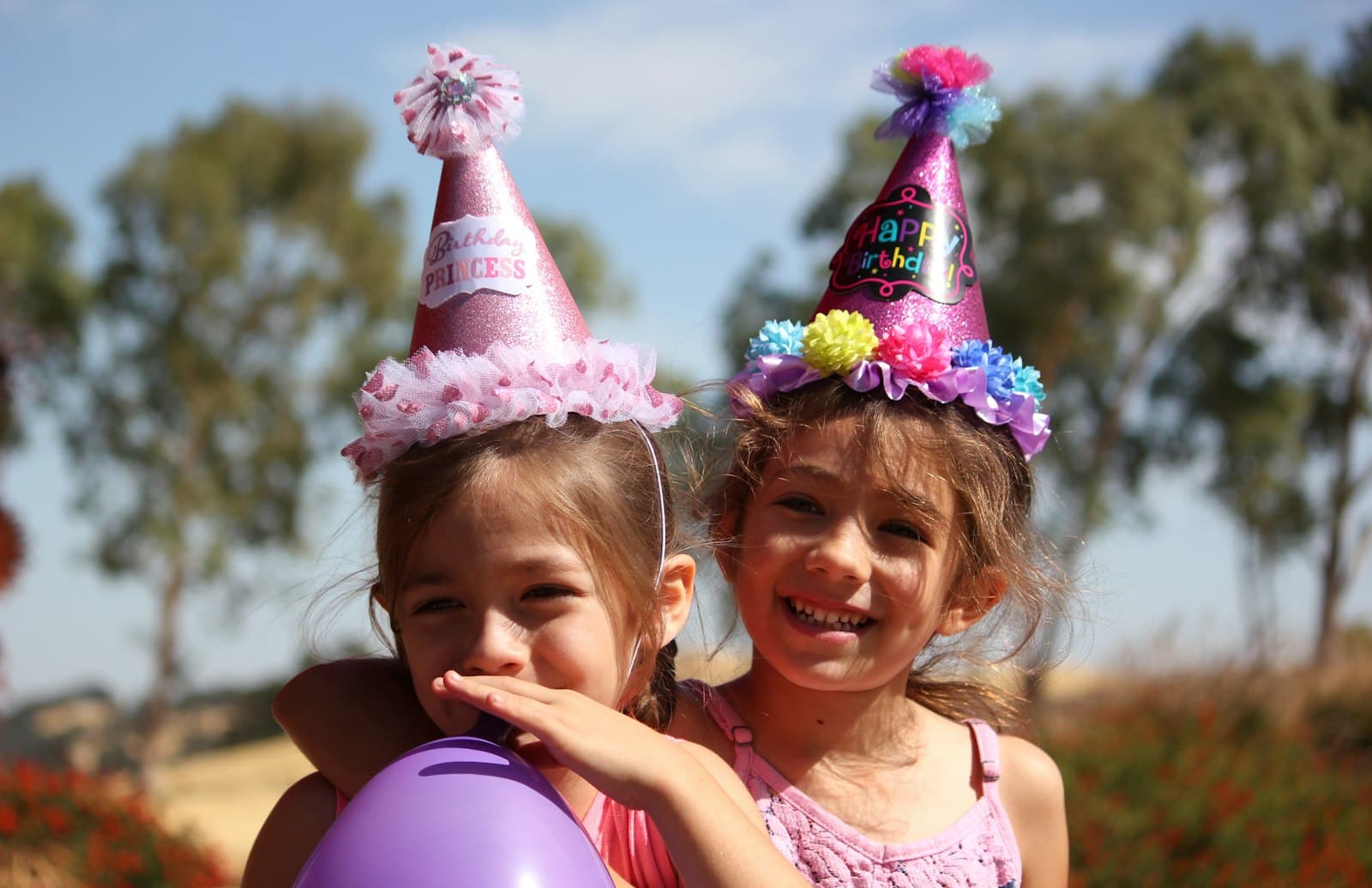 Dos nenas con gorros de cumpleaños y un globo violeta, riéndose al aire libre