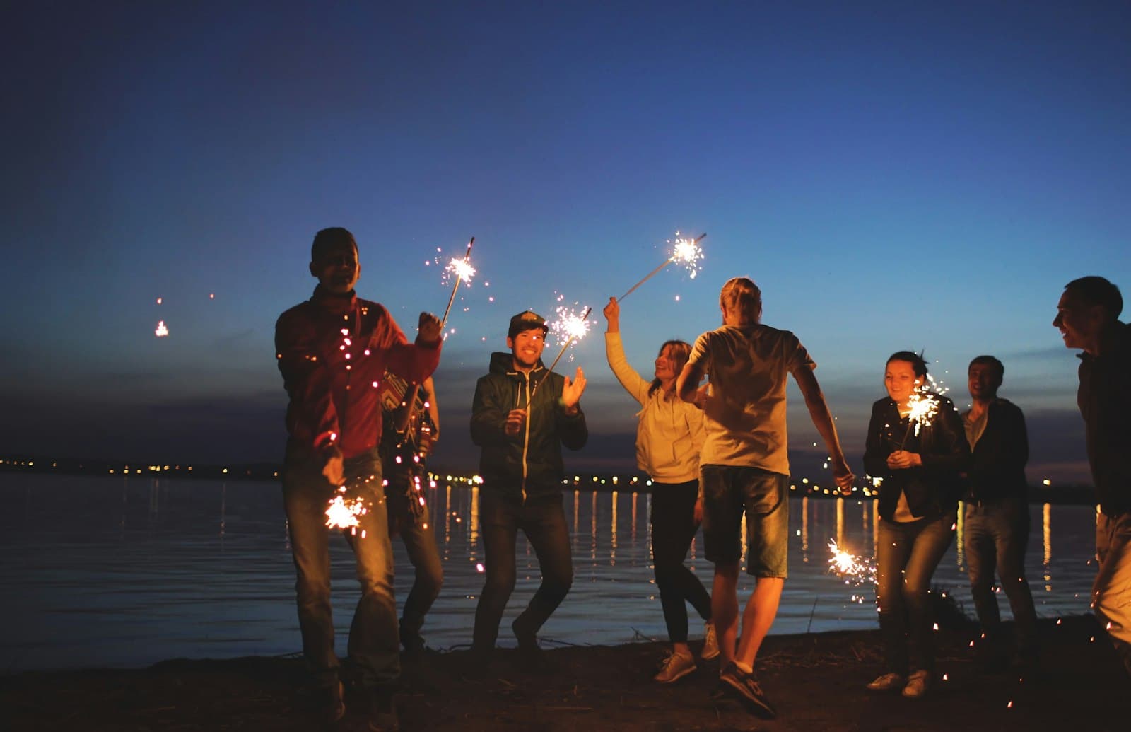 Grupo de amigos con bengalas encendidas de noche, festejando al lado del agua