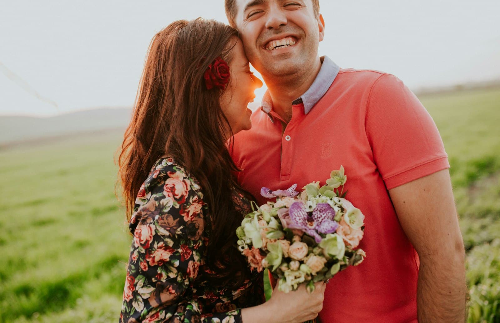 Pareja riéndose abrazada en un campo al atardecer, ella con un ramo de flores