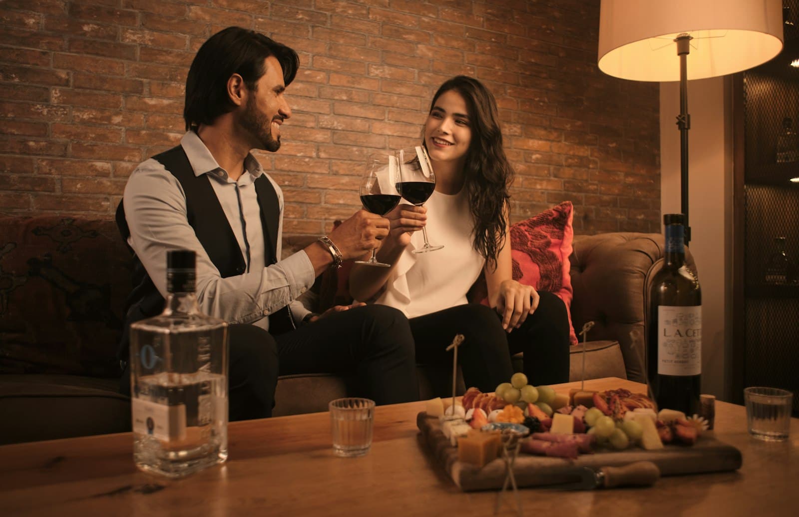 Pareja brindando con vino en el sillón de su casa, con una picada sobre la mesa