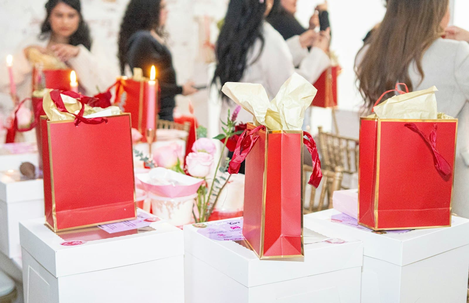 Mesa de baby shower con bolsas de regalo rojas y flores, con las invitadas charlando de fondo