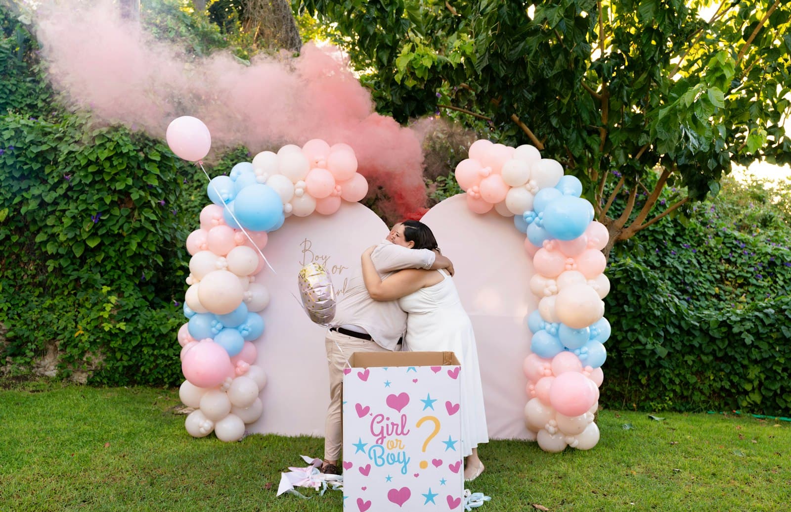 Pareja abrazándose frente a un arco de globos celestes y rosas, con humo rosa saliendo de una caja
